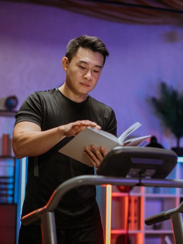 Attractive young sports asian man reading book while doing fitness exercise, running on treadmill